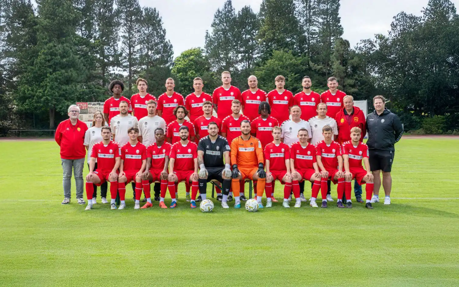 Fußballmannschaft in roten Trikots in drei Reihen auf dem Spielfeld, Trainer an den Seiten.